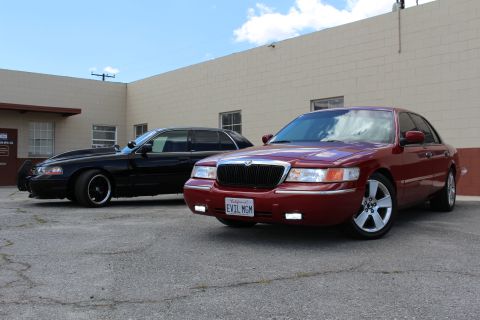 2003 Ford Crown Victoria - maroon Mercury Grand Marquis and a lowered black Ford sedan sit angled together before a tan warehouse wall.