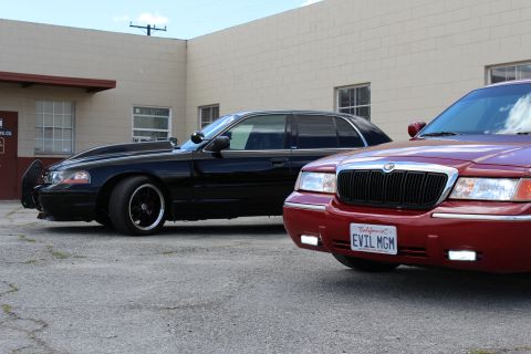 2003 Ford Crown Victoria - black Ford sedan parks beside the chrome-grilled front corner of a maroon Mercury bearing plate EVIL MGM.