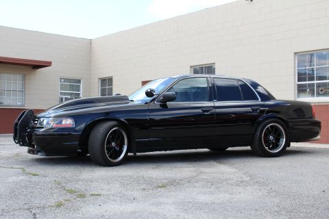 2003 Ford Crown Victoria - lowered black Ford Crown Victoria sits in side profile with a push bumper and dark wheels before a tan wall.