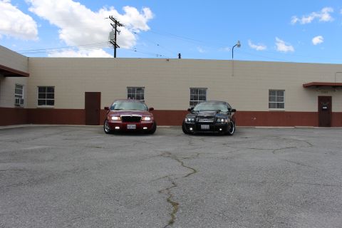 2003 Ford Crown Victoria - maroon Mercury sedan and a black Ford sedan park side by side facing the camera before a tan warehouse.
