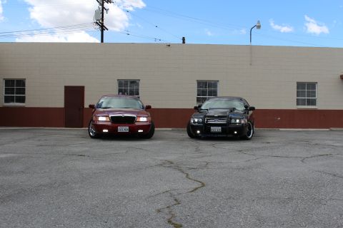 2003 Ford Crown Victoria - maroon Mercury and a lowered black Ford sedan sit head-on together against a tan building with paned windows.
