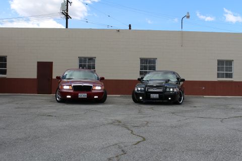 2003 Ford Crown Victoria - maroon Mercury sedan and a black Ford sedan with a police push bumper face the camera in a sunny parking lot.