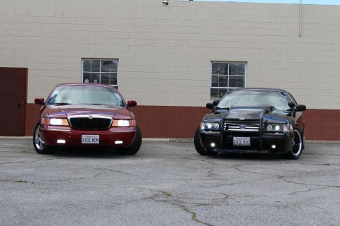 2003 Ford Crown Victoria - maroon Mercury Grand Marquis and a black Crown Victoria with bull bar park nose-out before a tan industrial wall.