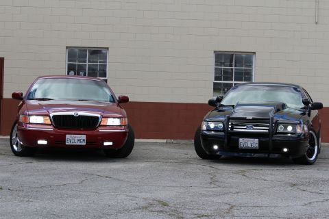 2003 Ford Crown Victoria - maroon Grand Marquis and black Crown Victoria with chrome wheels and grille guard sit side by side on cracked asphalt.