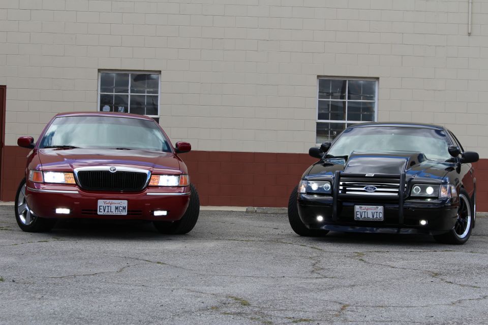 Maroon Mercury sedan and black Ford Crown Victoria with bull bar parked outside tan building