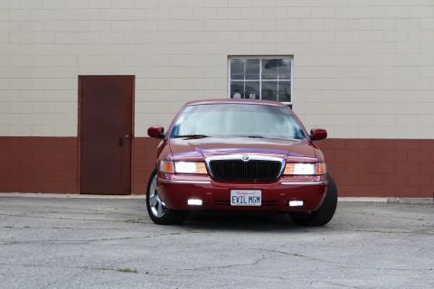 2003 Ford Crown Victoria - Front view of a lowered maroon Mercury Grand Marquis with chrome wheels parked on cracked pavement by a tan wall.