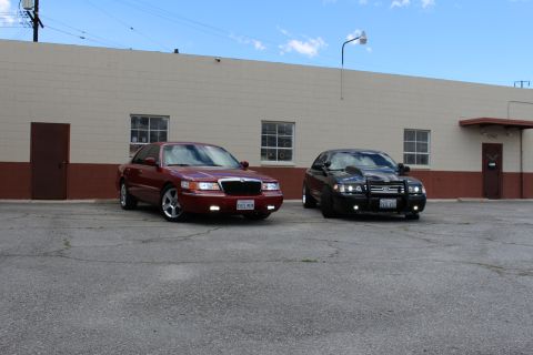 2003 Ford Crown Victoria - maroon Grand Marquis and a black Crown Victoria with grille guard park side by side beneath a clear blue sky.