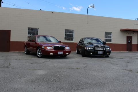 2003 Ford Crown Victoria - Front three-quarter view of a maroon Mercury sedan beside a black Crown Victoria with bull bar in a sunlit lot.