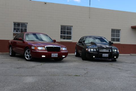 2003 Ford Crown Victoria - maroon Grand Marquis with chrome wheels parks beside a black Crown Victoria wearing a bull bar at a tan building.