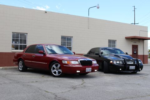 2003 Ford Crown Victoria - maroon Mercury Grand Marquis angles toward the camera with a darker Ford sedan parked behind it near a tan wall.