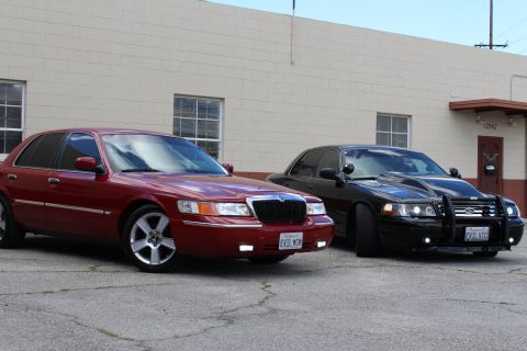 2003 Ford Crown Victoria - maroon Mercury Grand Marquis sits in front of a black Crown Victoria with a chrome bull bar near a tan building.