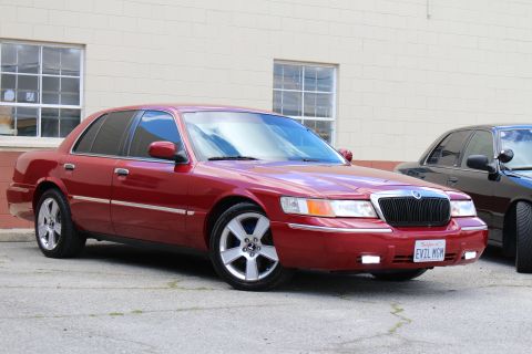 2003 Ford Crown Victoria - lowered maroon Mercury Grand Marquis with chrome wheels and EVIL MGM plate parks beside another dark car at a tan wall.
