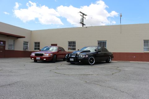 2003 Ford Crown Victoria - maroon Grand Marquis and a black Crown Victoria with grille guard sit in a lot under a cloud-streaked blue sky.