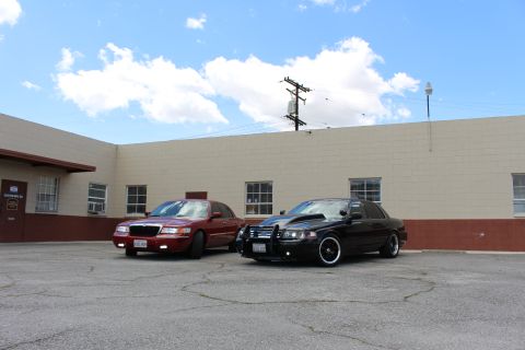2003 Ford Crown Victoria - Wide view of a maroon Grand Marquis beside a black Crown Victoria with push bar in a paved lot under partly cloudy sky.