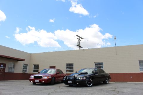 2003 Ford Crown Victoria - maroon Grand Marquis and a black Crown Victoria with grille guard park side by side under a bright cloudy sky.