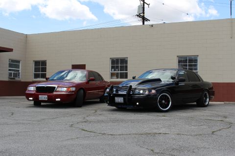 2003 Ford Crown Victoria - dark red Mercury sedan and a black Ford Crown Victoria with a black push bar are parked together on cracked asphalt.