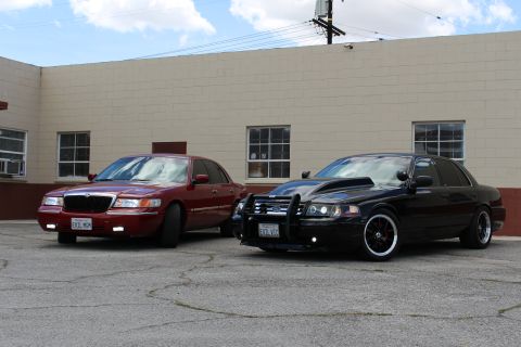 2003 Ford Crown Victoria - maroon Mercury sedan and a lowered black Ford Crown Victoria with a push bar are parked nose-out in front of a tan building.