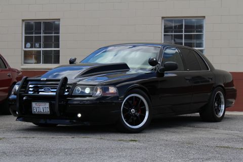 2003 Ford Crown Victoria - lowered black Ford Crown Victoria with a black push bar and black multi-spoke wheels sits in front of a tan building.