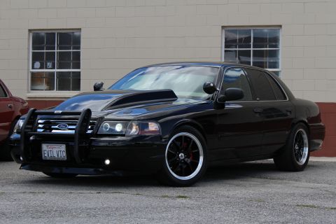 2003 Ford Crown Victoria - lowered black Ford Crown Victoria with a push bar and California EVIL VIC plate is parked beside a brick-trimmed wall.