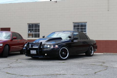 2003 Ford Crown Victoria - Front three-quarter view of a lowered black Ford Crown Victoria with a push bar and black wheels, a red sedan partly visible at left.
