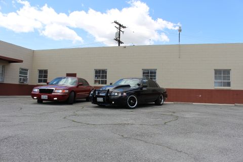 2003 Ford Crown Victoria - dark red Mercury sedan and a black Ford Crown Victoria with a push bar are parked together under utility wires and a bright sky.