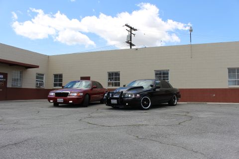 2003 Ford Crown Victoria - maroon Mercury sedan and a black Ford Crown Victoria sit side by side in front of a long tan building under a blue sky.