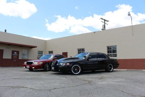 2003 Ford Crown Victoria - dark red Mercury sedan and a black Ford Crown Victoria with a push bar are angled in a parking lot in front of a tan building.