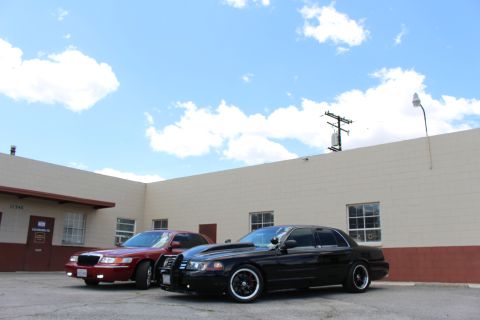 2003 Ford Crown Victoria - maroon Mercury sedan and a black Ford Crown Victoria are parked together below a tall blue sky with scattered clouds.