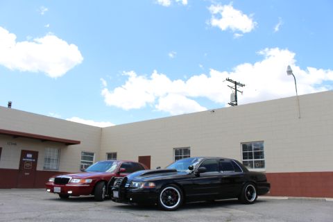 2003 Ford Crown Victoria - dark red Mercury sedan and a black Ford Crown Victoria sit side by side near a tan wall beneath a wide blue sky.