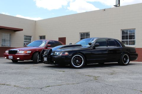 2003 Ford Crown Victoria - red Mercury sedan parked beside a lowered black Ford Crown Victoria with a push bar and black wheels in front of a tan building.