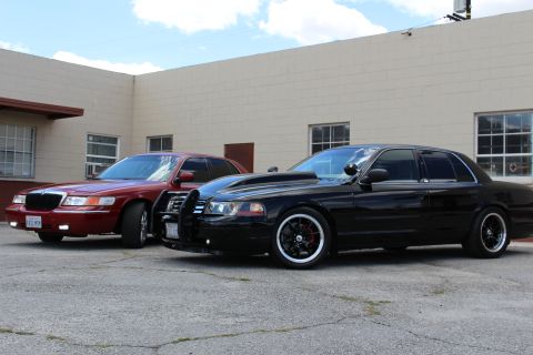 2003 Ford Crown Victoria - dark red Mercury sedan sits beside a lowered black Ford Crown Victoria with a push bar and whitewall-style tires.