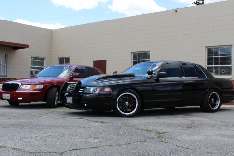 2003 Ford Crown Victoria - maroon Mercury sedan and a lowered black Ford Crown Victoria with a push bar are parked together by a tan building.