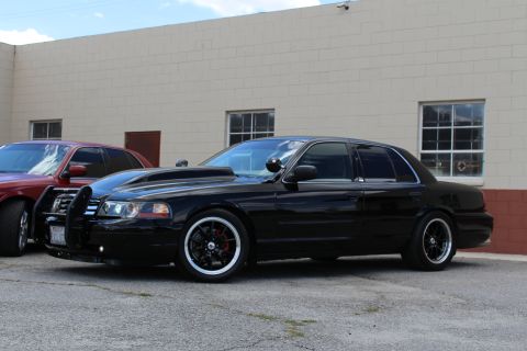 2003 Ford Crown Victoria - lowered black Ford Crown Victoria with a push bar and black wheels parked in front of a tan wall, a red sedan visible at left.