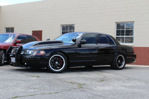 2003 Ford Crown Victoria - lowered black Ford Crown Victoria with a push bar and black wheels sits angled by a tan building, a red sedan partly visible.