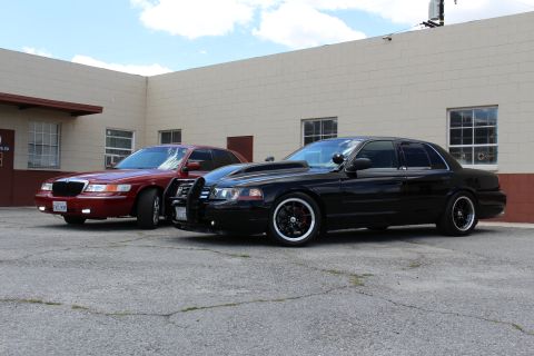 2003 Ford Crown Victoria - maroon Mercury sedan and a lowered black Ford Crown Victoria with a push bar are parked together near a brick-trimmed wall.