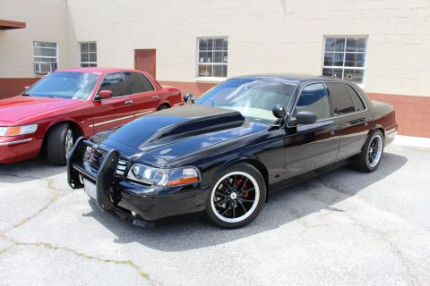 2003 Ford Crown Victoria - red Mercury sedan and a lowered black Ford Crown Victoria with a push bar and black wheels are parked by a tan building.