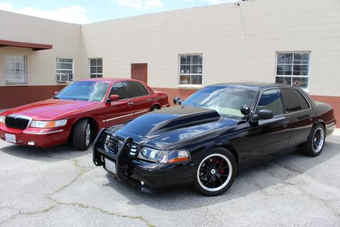 2003 Ford Crown Victoria - red Mercury sedan and a lowered black Ford Crown Victoria with a push bar sit side by side on cracked asphalt by a tan building.