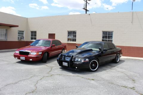 2003 Ford Crown Victoria - maroon Mercury sedan and a lowered black Ford Crown Victoria with a push bar are parked together under utility wires and clouds.