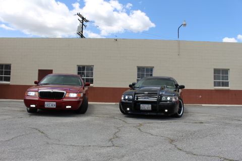 2003 Ford Crown Victoria - Front view of a dark red Mercury sedan (EVIL MGM) and a black Ford with a push bar (EVIL VIC) parked side by side under a blue sky.