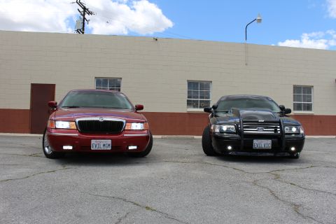 2003 Ford Crown Victoria - maroon Mercury sedan beside a black Crown Victoria with a brush guard, both facing the camera in a cracked lot.