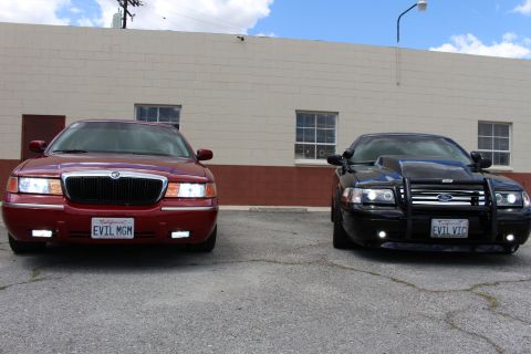 2003 Ford Crown Victoria - dark red Mercury sedan beside a black Ford Crown Victoria with a brush guard, both facing the camera in a lot.