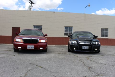 2003 Ford Crown Victoria - maroon Mercury sedan and a black Crown Victoria with a grille guard sit side by side outside a tan building.