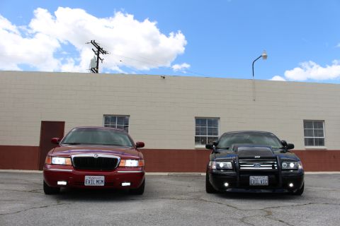 2003 Ford Crown Victoria - dark red Mercury sedan and a black Ford Crown Victoria parked side by side in a lot beneath a blue, cloud-filled sky.