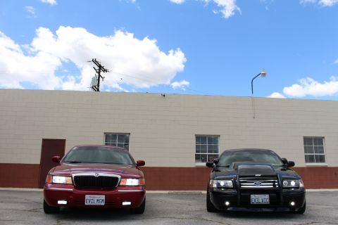 2003 Ford Crown Victoria - maroon Mercury sedan and a black Crown Victoria with a brush guard face the camera outside a long tan building.