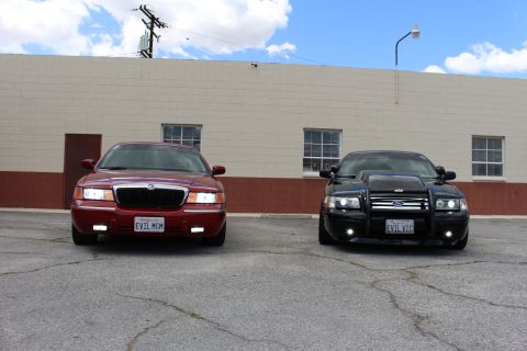 2003 Ford Crown Victoria - dark red Mercury sedan and a black Ford Crown Victoria with a grille guard sit side by side in a cracked parking lot.