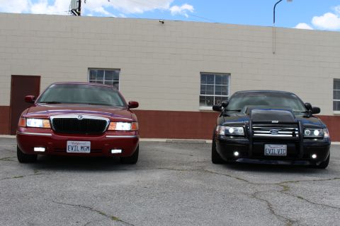 2003 Ford Crown Victoria - maroon Mercury sedan beside a black Crown Victoria with a brush guard, both photographed head-on in a lot.