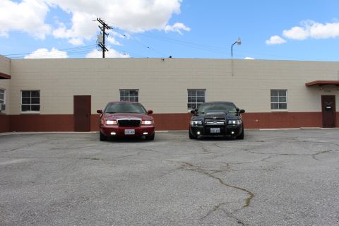 2003 Ford Crown Victoria - maroon Mercury sedan and a black Ford sedan parked side by side, seen at a distance in front of a long tan wall.