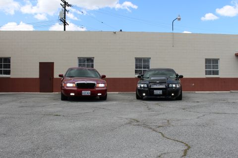 2003 Ford Crown Victoria - Front view of a maroon Mercury sedan and a black sedan parked side by side on cracked asphalt before a tan warehouse wall.