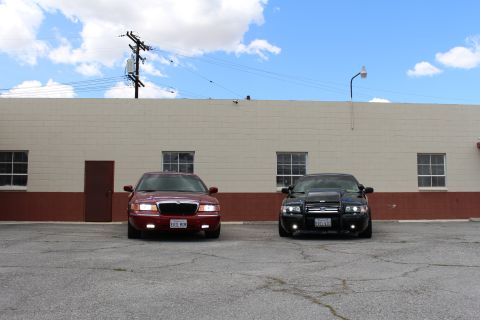2003 Ford Crown Victoria - maroon Mercury and a black sedan sit nose-forward in a lot under a blue sky with a power pole behind the building.