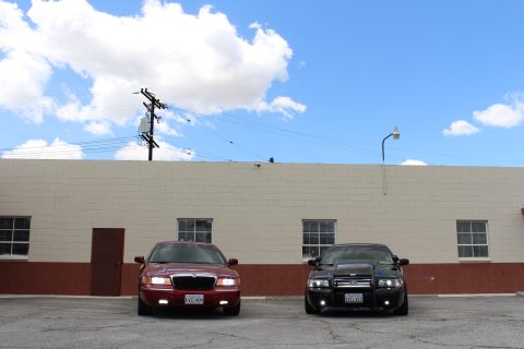 2003 Ford Crown Victoria - Wide shot of a maroon Mercury and a black sedan parked front-facing against a long warehouse under puffy clouds.